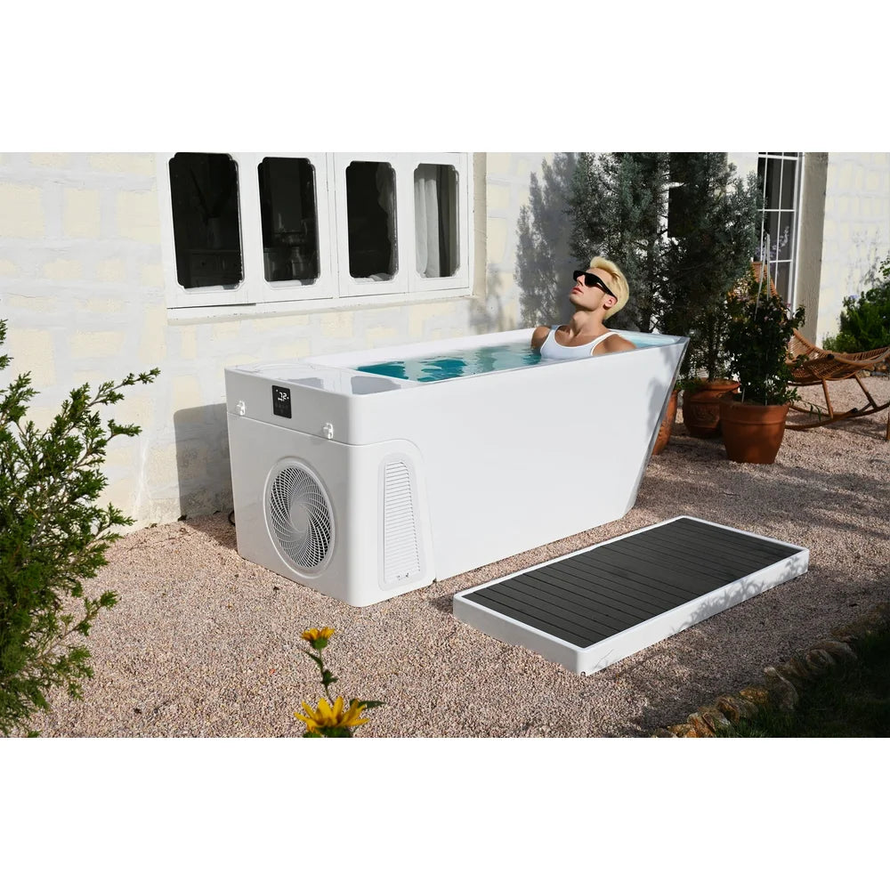 Person reclining in the AMBOHR All-in Cold Plunge Spa on a gravel courtyard patio beside a stone house