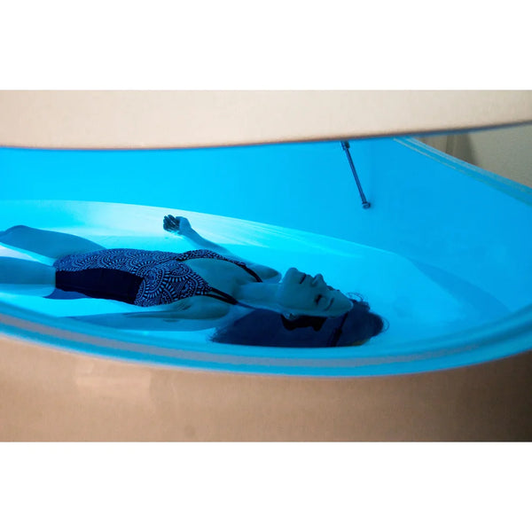Woman floating full-length inside a turquoise-lit Dreampod V2 with the lid held open by a gas spring strut