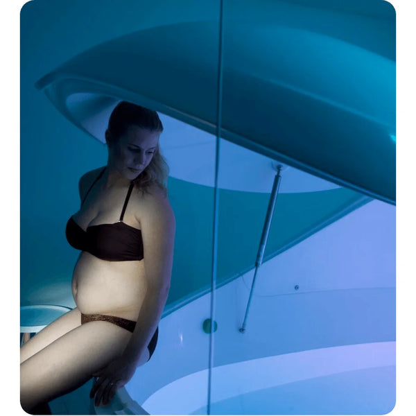 Pregnant woman sitting on the rim of a Dreampod V2 Float Pod under the raised lid in teal-blue light