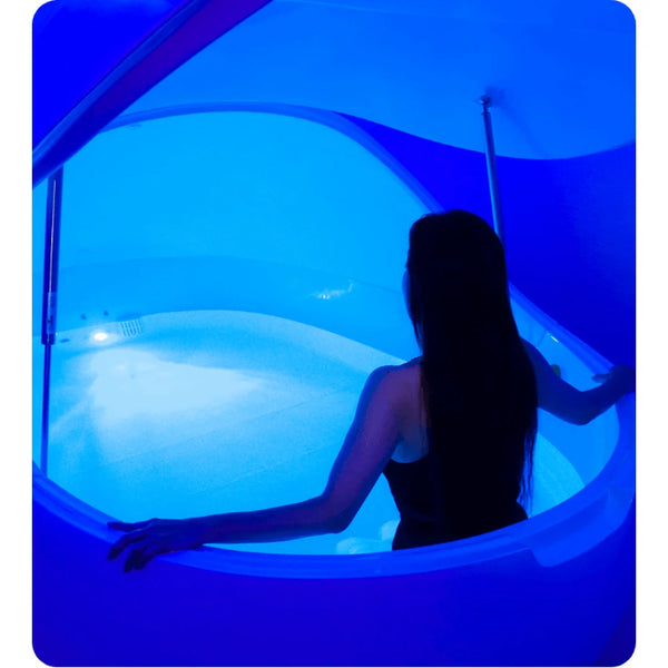 Woman stepping into an open Dreampod V2 Isolation Tank bathed in deep blue light above shallow salt water