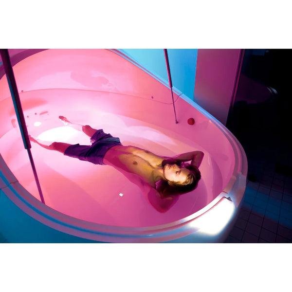 High angle of a man floating in pink lit salt water showing the moulded shell and fittings of the Dreampod Vmax Isolation Pod