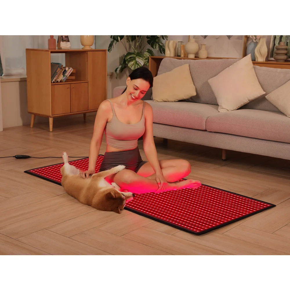 Smiling woman pets her dog while seated on the lit floor mat of the Kore KoreKapsule Red Light Therapy Chamber Kit