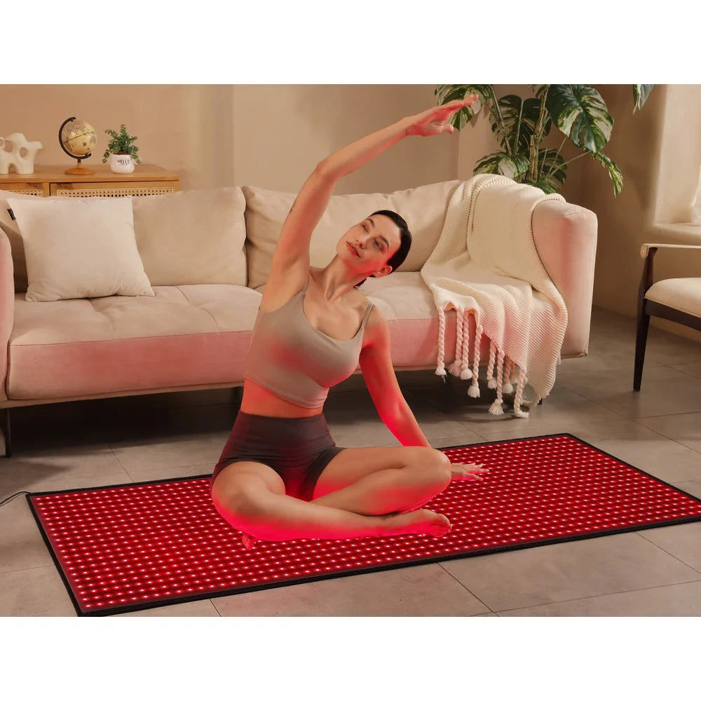 Woman stretching cross-legged on the lit pad beside a sofa, using Kore KoreKapsule Red Light Therapy Devices at home