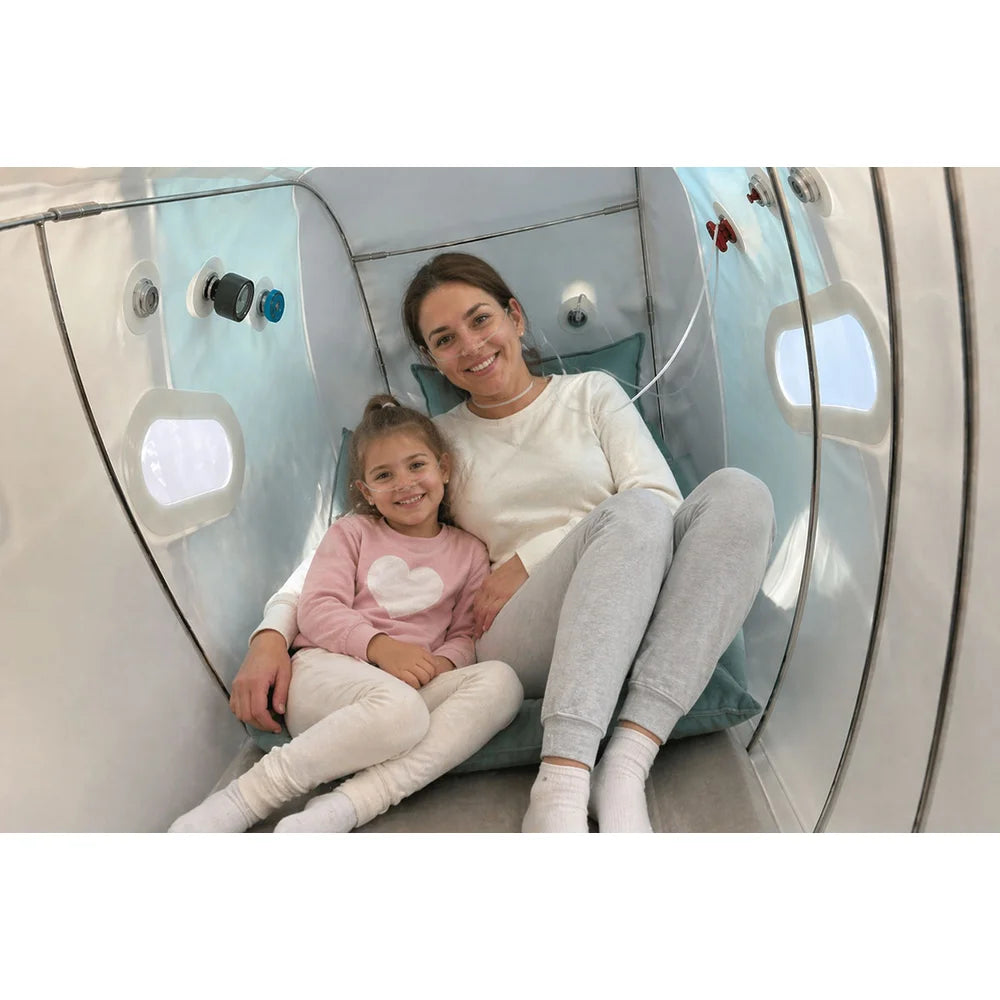 Smiling woman and young girl wearing nasal cannulas inside the Oxyrevo Forward90 Sitting Hyperbaric Chamber, teal cushions and lit windows behind them