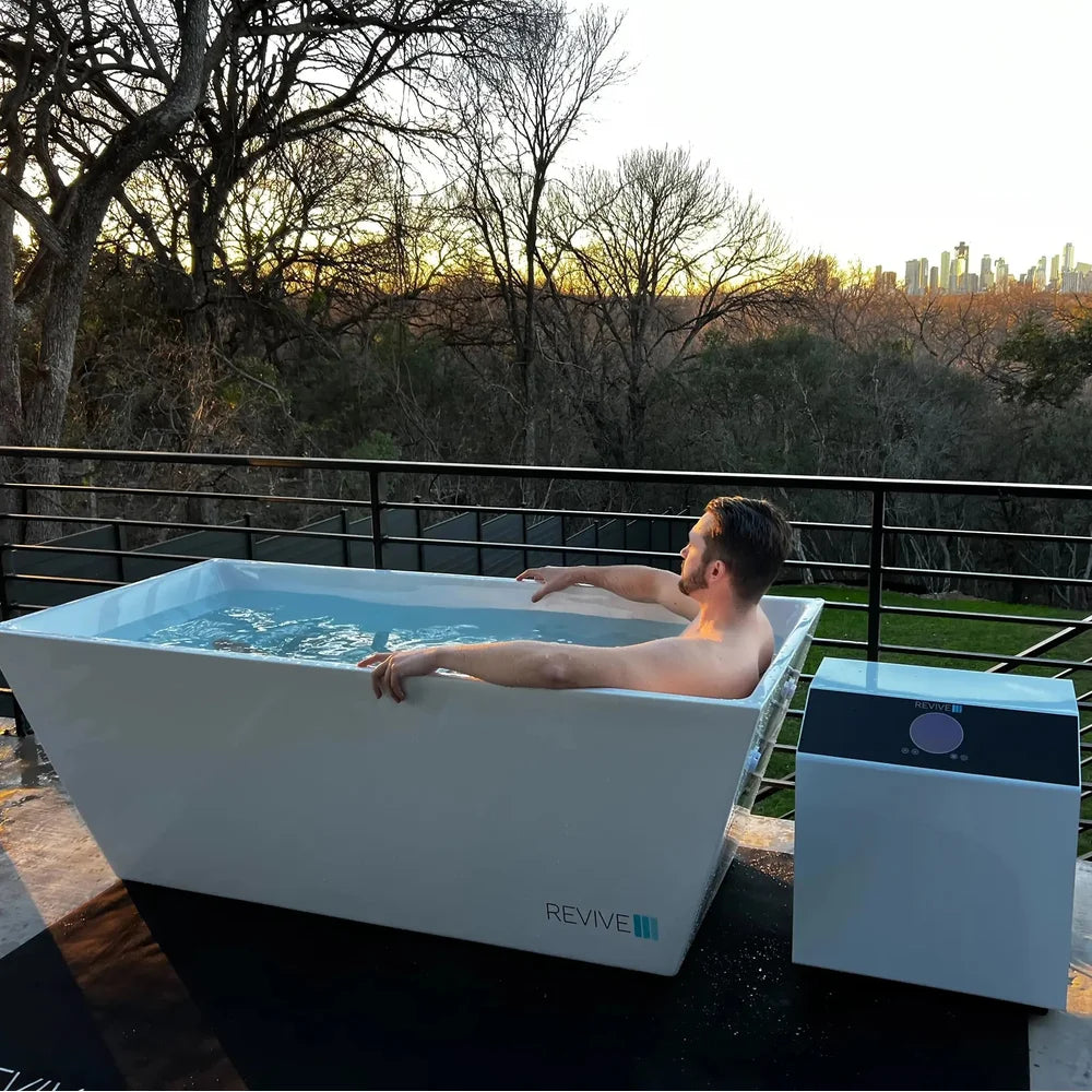 Man soaking in the Revive Plunge Acrylic Plunge Tub on a deck at sunset with the chiller alongside