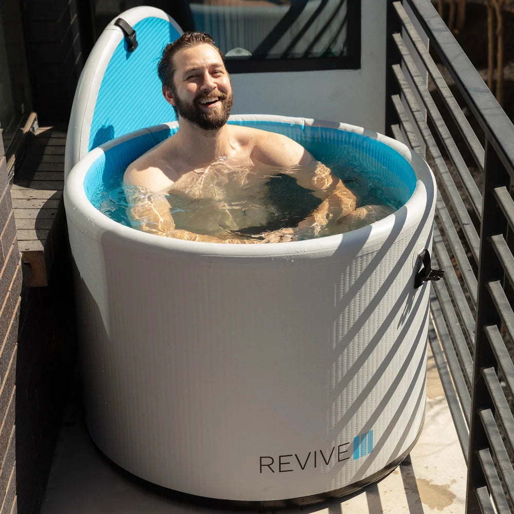 Smiling man soaking in the Revive Inflatable Pod Plunge Ice Bath Tub set up in a narrow outdoor walkway