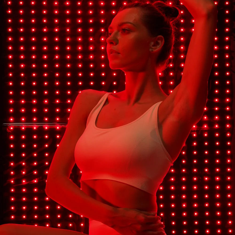 Woman in a white sports top poses before glowing red LED rows of the Kore KoreKapsule Near Infrared Light Therapy System