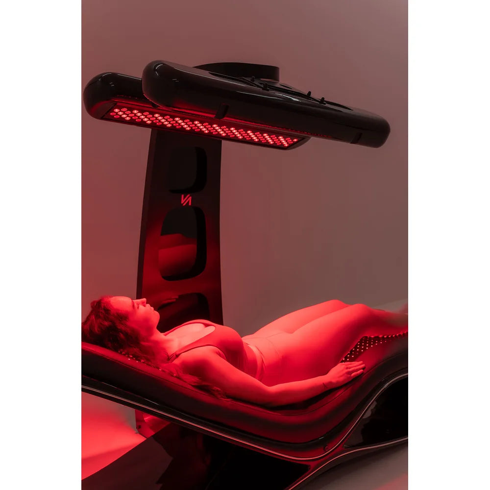 Client relaxing beneath the illuminated overhead lamp of the Vacu Activ Red Couch 360 Light Therapy Bed in a studio